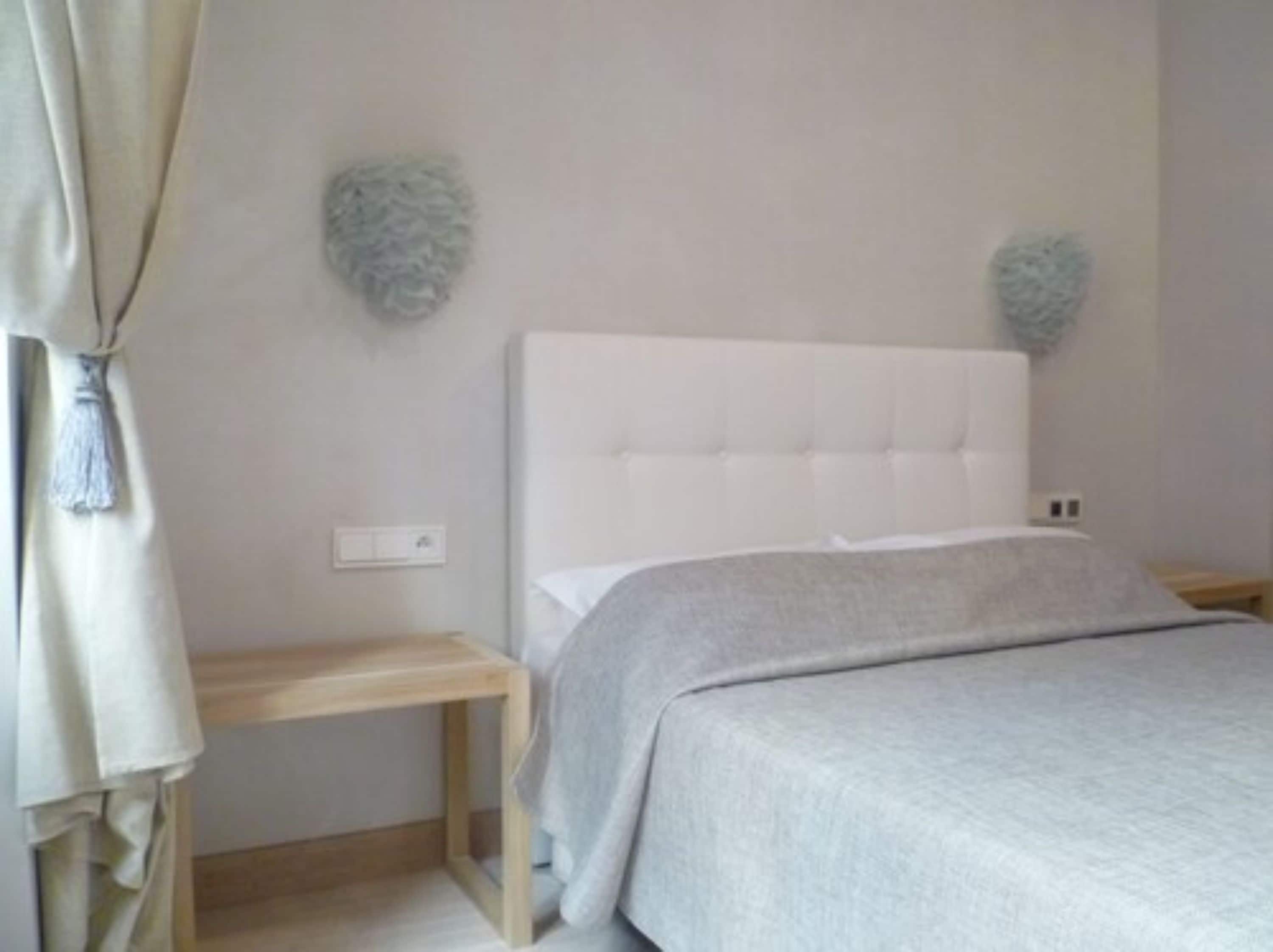 Quarto com uma cama branca, uma mesinha de cabeceira de madeira e um interruptor de luz fixado na parede.