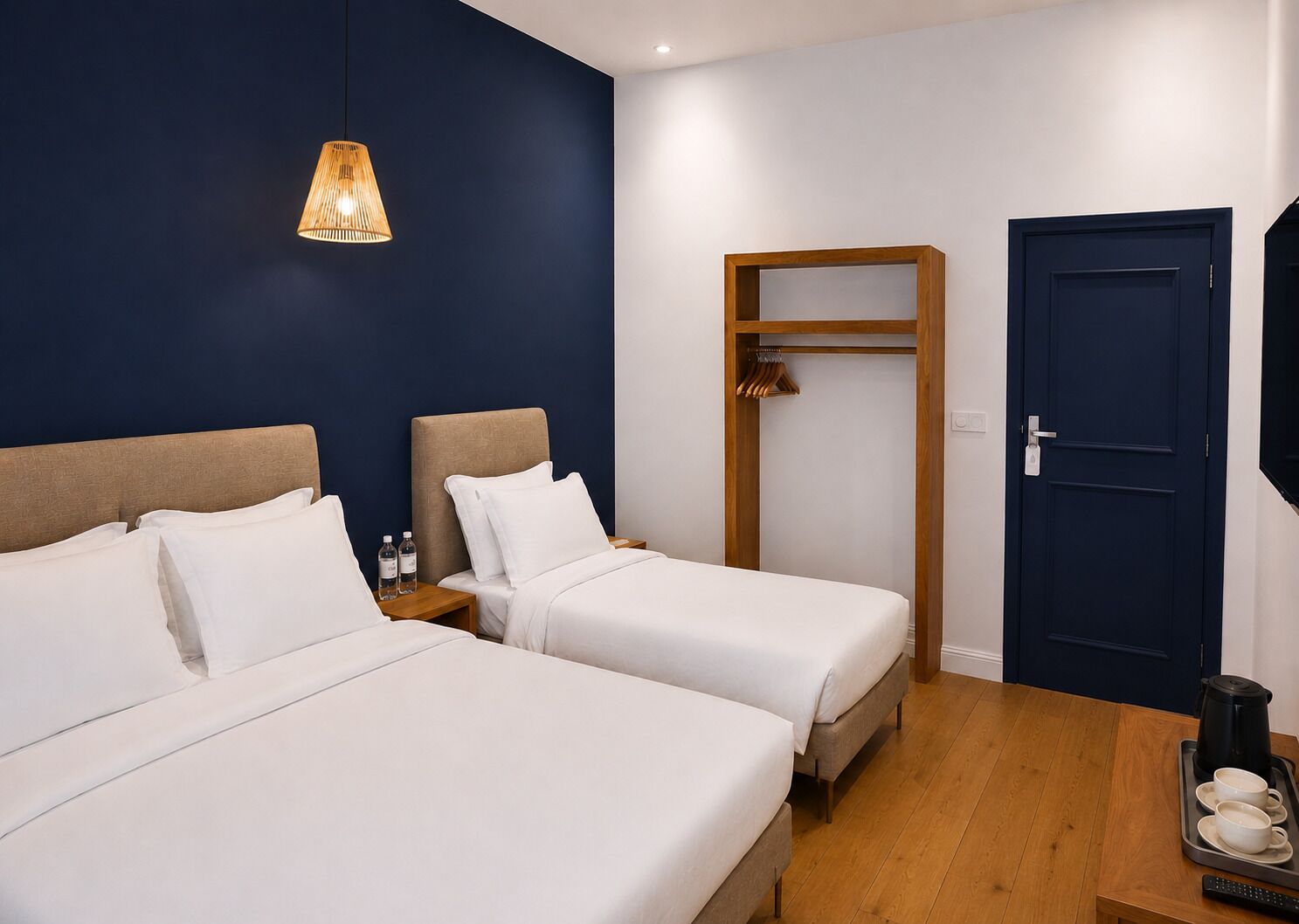 Una camera d'albergo con due letti, un armadio in legno, una porta blu scuro e una lampada a sospensione.