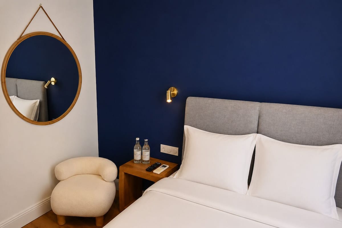 Une chambre d’hôtel avec un lit, un miroir rond, une table de chevet avec deux bouteilles, une télécommande et une lampe fixée au mur.