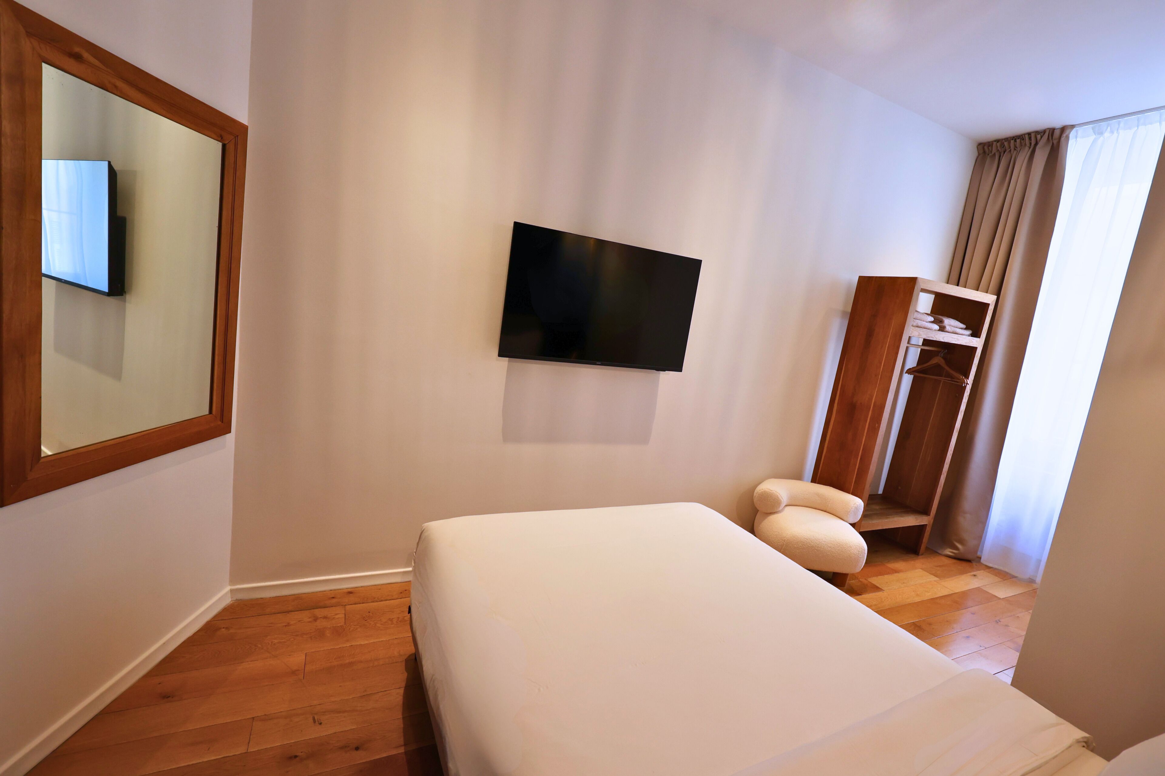 A hotel room with a bed, a TV mounted on the wall, a wooden wardrobe, and a window with curtains.