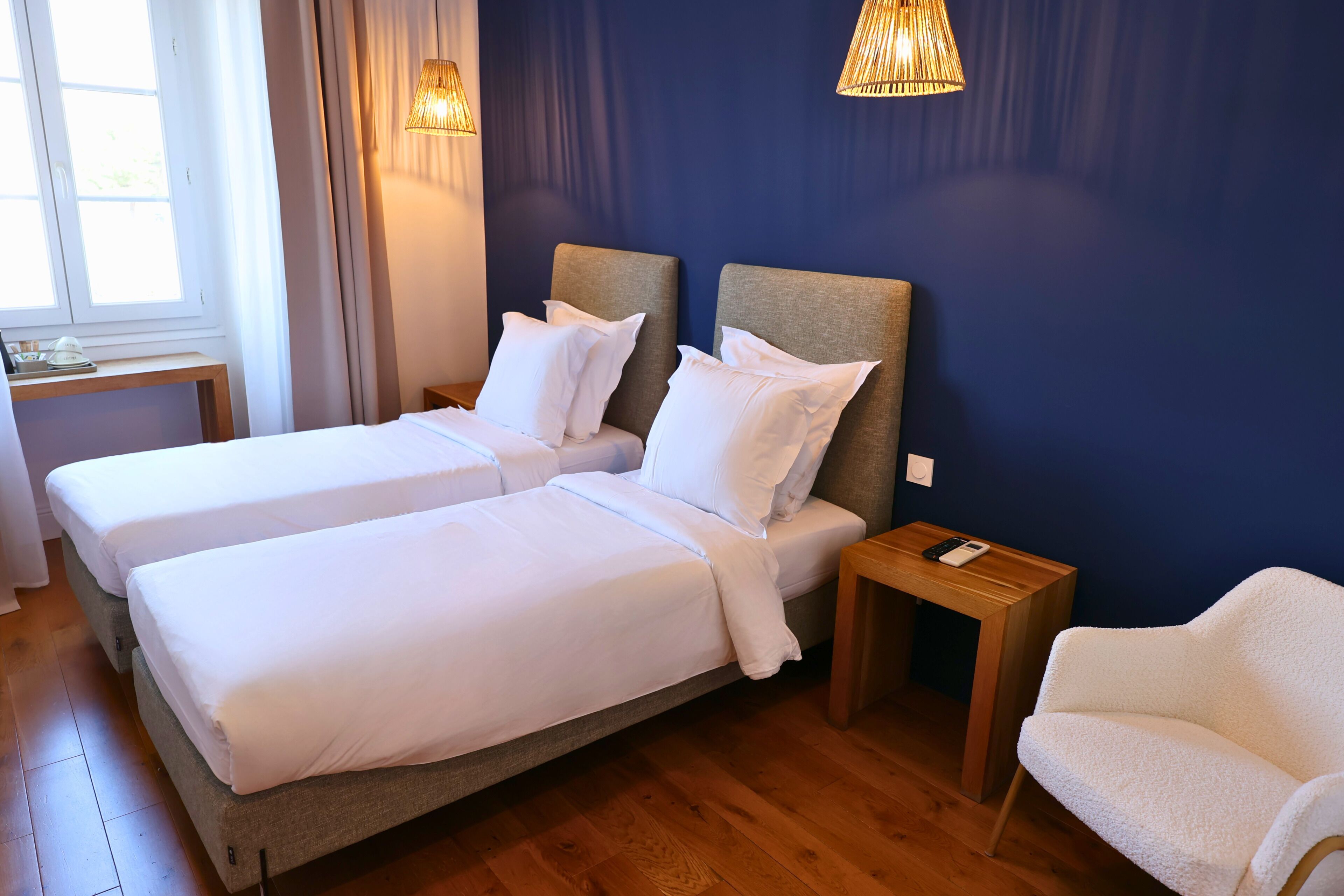 Een hotelkamer met twee bedden, houten vloeren, een nachtkastje met een lamp, een stoel en een raam met gordijnen.