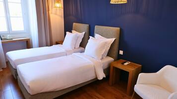 Een hotelkamer met twee bedden, houten vloeren, een nachtkastje met een lamp, een stoel en een raam met gordijnen.