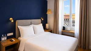 Una habitación de hotel con cama, mesitas de noche, lámpara, ventana con cortinas y vista a un paisaje urbano.