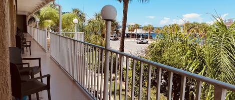 A balcony with chairs, a railing, and a view of palm trees and a road with vehicles.