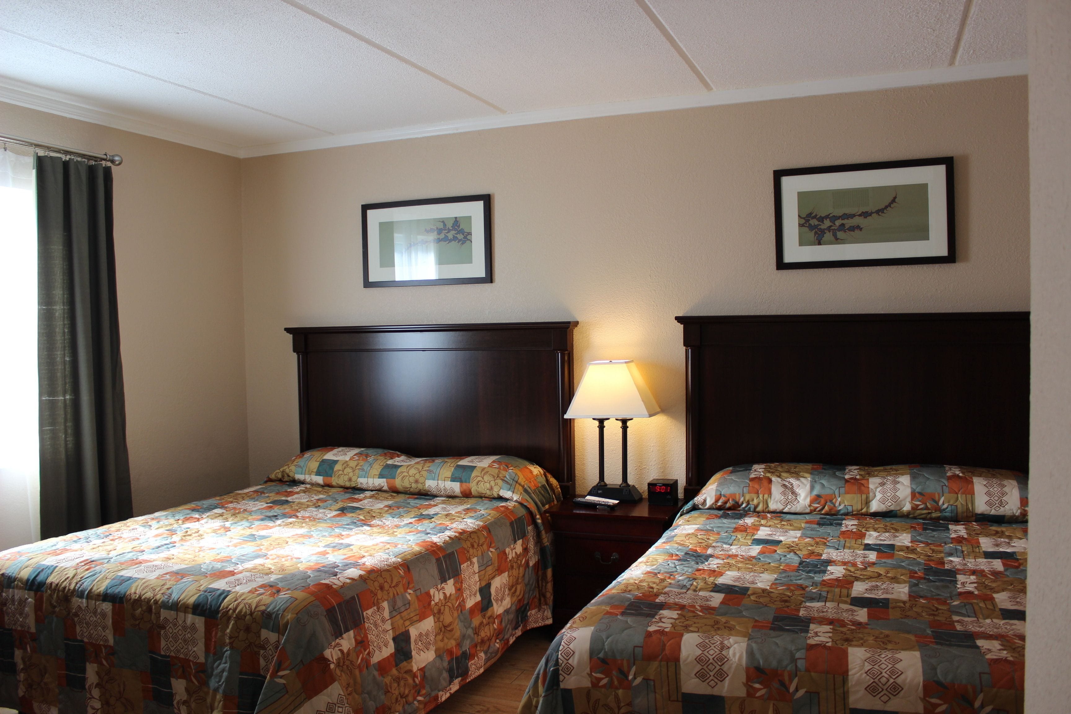 A hotel room with two beds, a nightstand, a lamp, and two framed pictures on the wall.