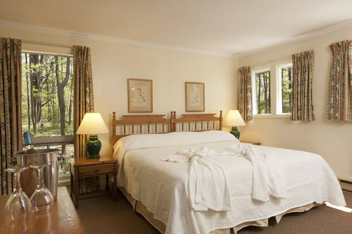 A bedroom with a large bed, two bedside tables with lamps, framed artwork on the wall, and a window with curtains.