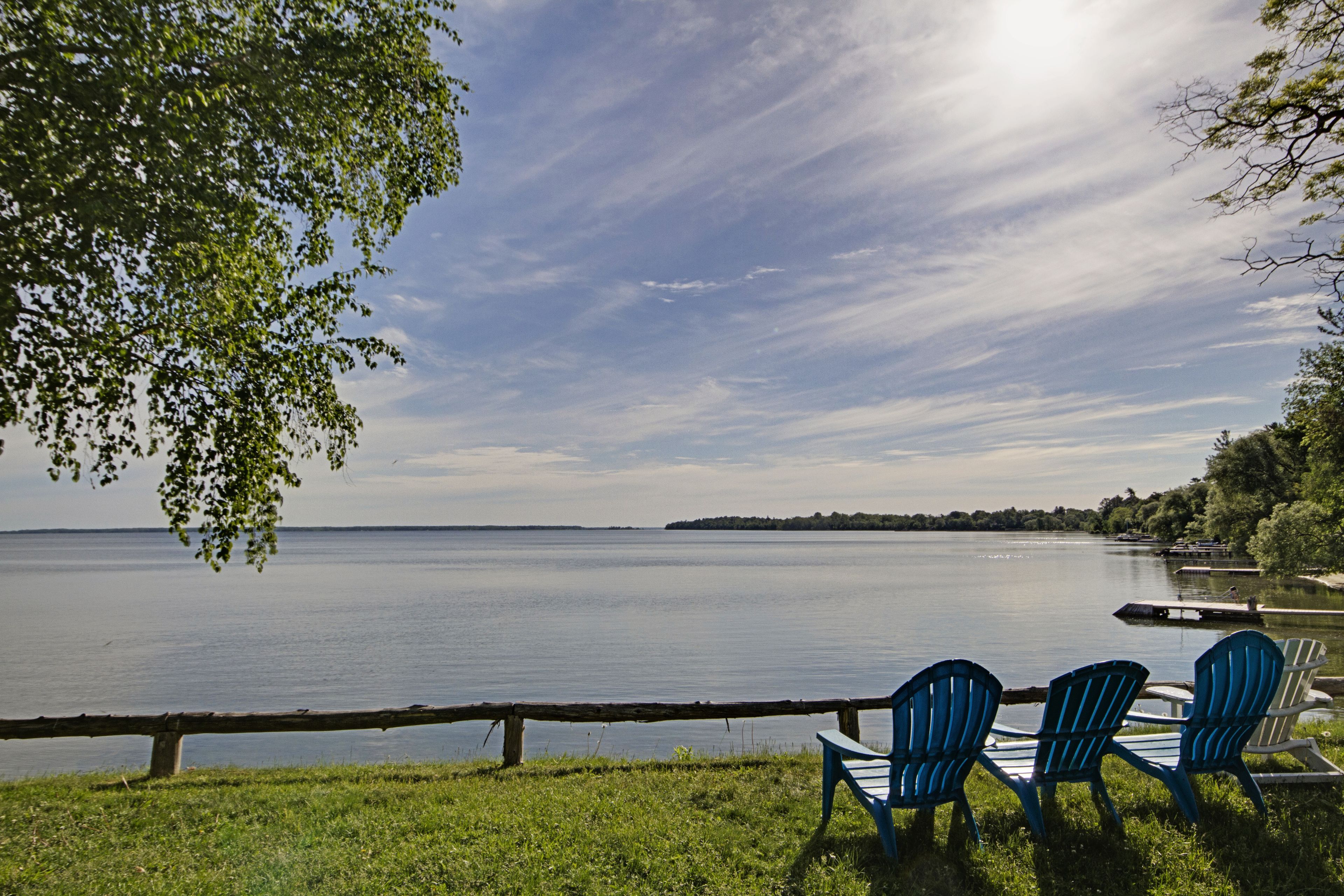 A lakeside view with a wooden fence, three Adirondack chairs, and a distant shoreline.