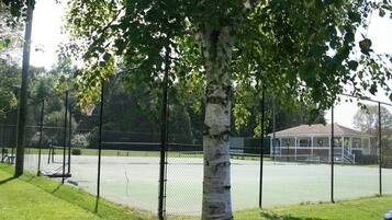 A tennis court enclosed by a chain-link fence, with a tree in the foreground and a building in the background.