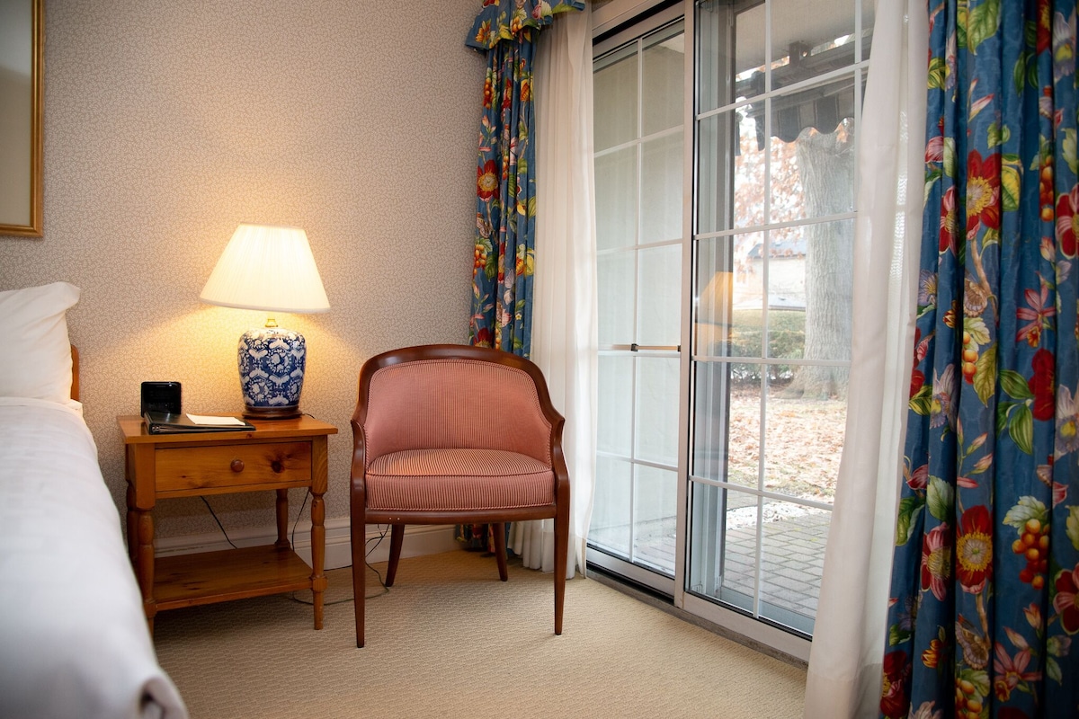 Habitación de hotel con cama, mesita de noche con lámpara, una silla y vistas a través de una puerta de vidrio.