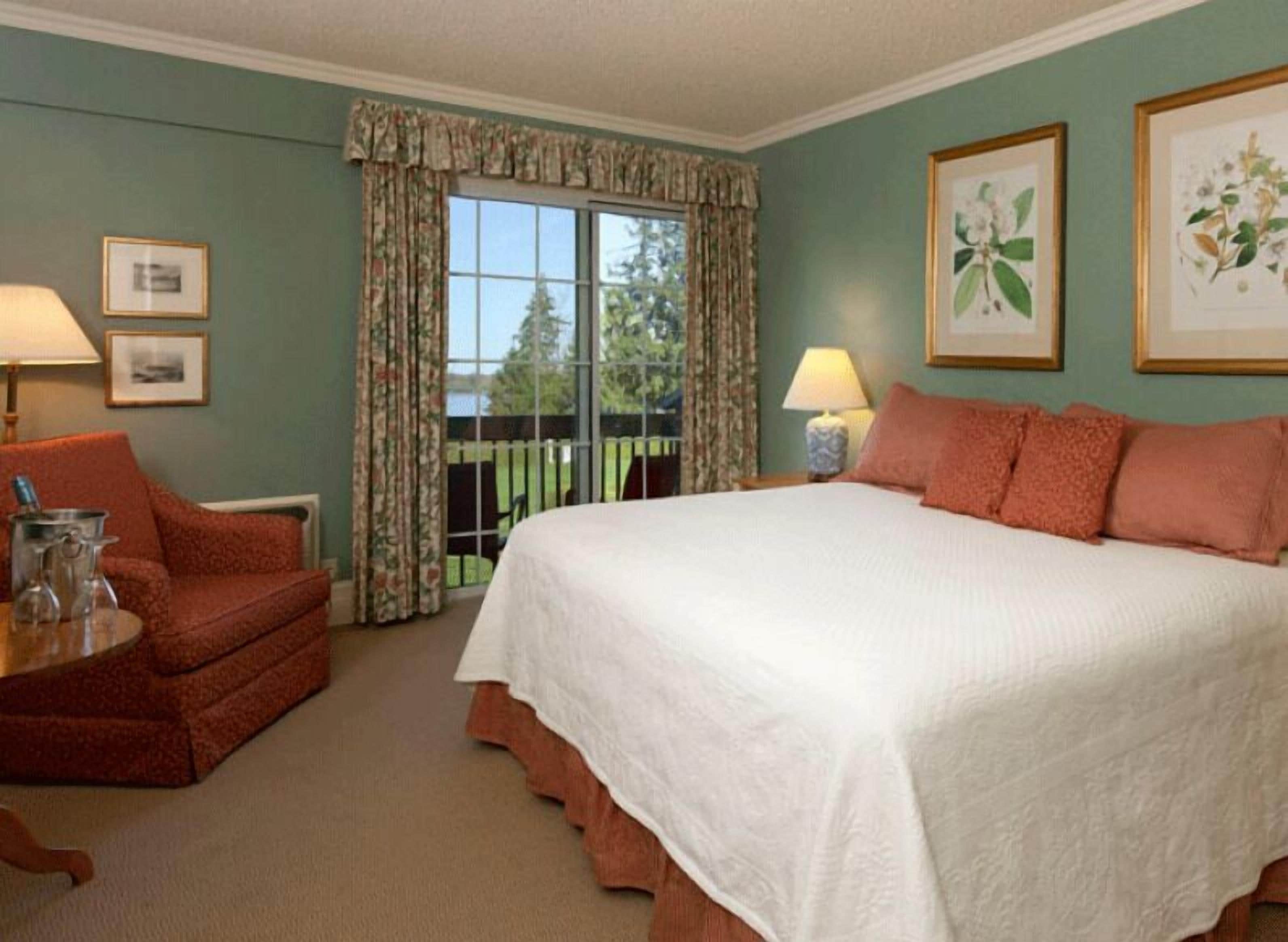 Une chambre à coucher avec un grand lit, une chaise, une table avec une lampe et une fenêtre avec des rideaux.