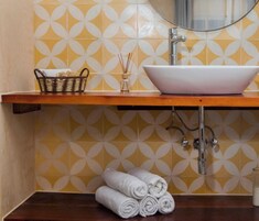 Un baño con un lavamanos blanco tipo lavabo, un estante de madera, una cesta de mimbre, una botella de vidrio con ramitas y toallas enrolladas sobre una superficie de madera oscura.