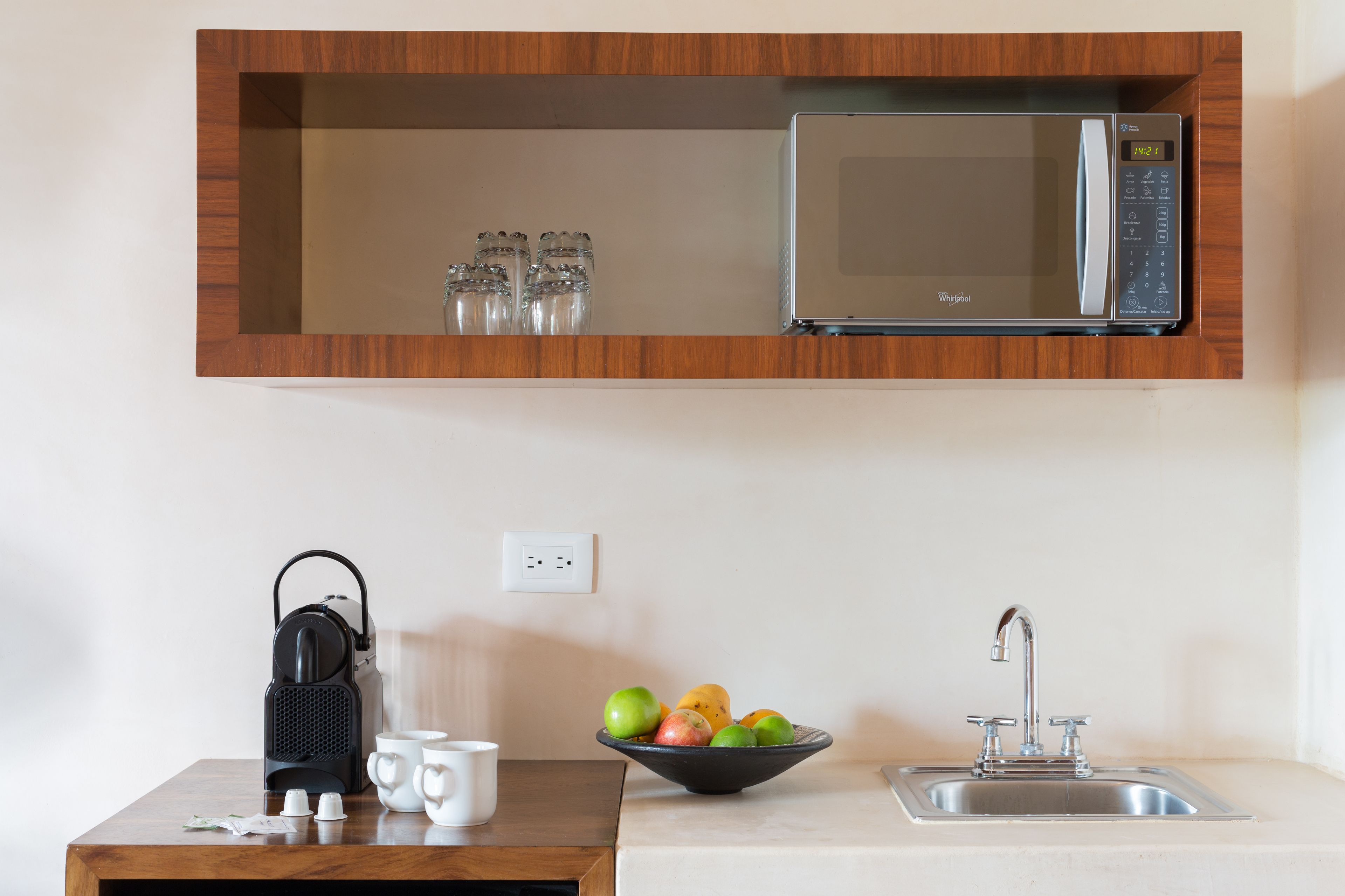 A kitchen with a microwave, a sink, a bowl of fruit, and a kettle on a wooden table.