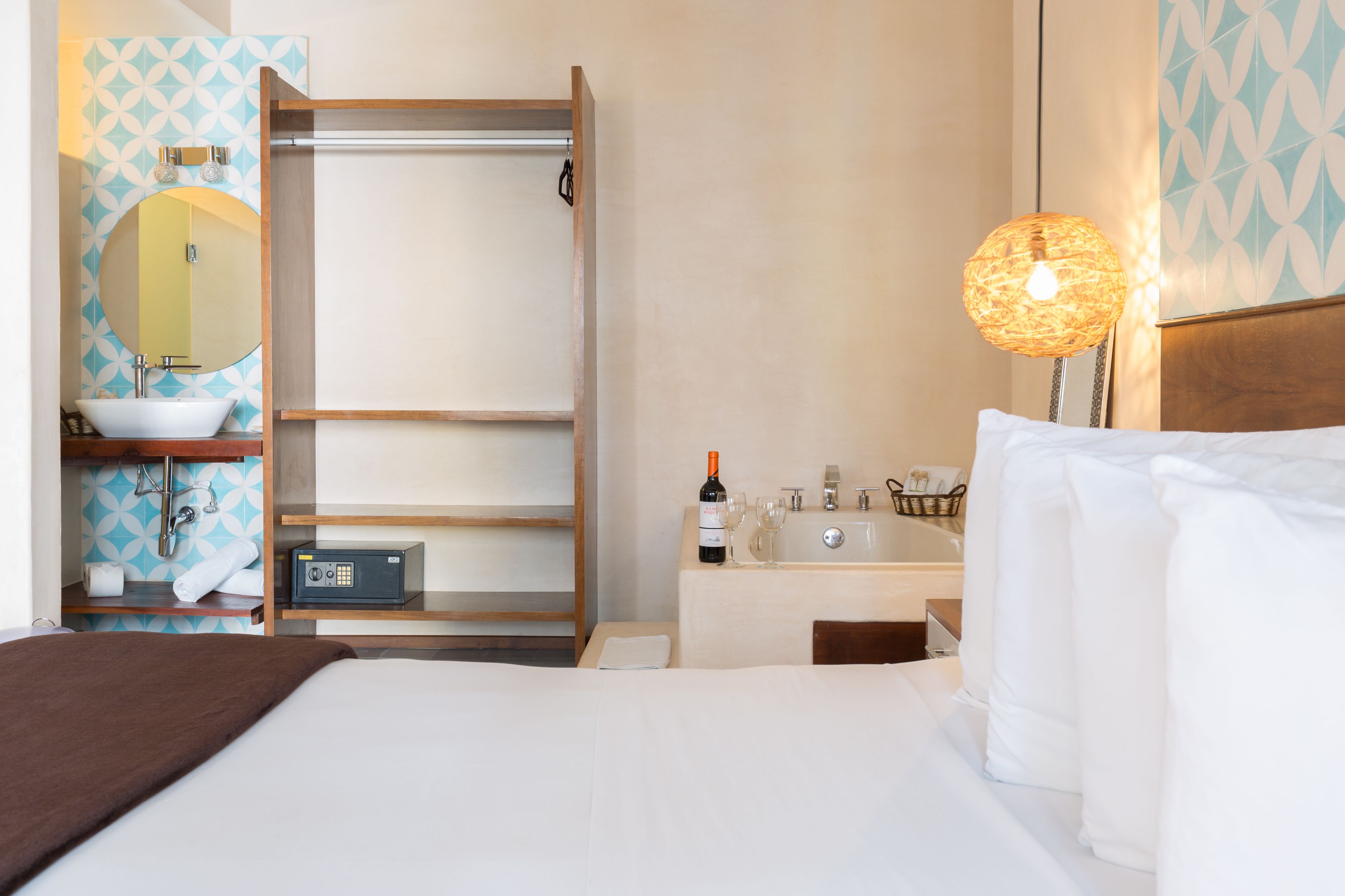 A hotel room with a bed, a bedside table with a bottle and glasses, a small desk with a lamp, a wardrobe, and a bathroom with a sink and mirror.
