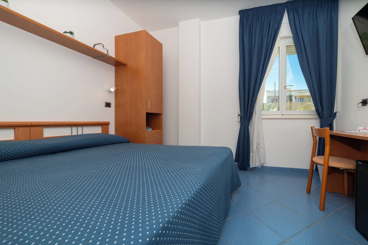 A bedroom with a bed, a wooden wardrobe, a desk with a chair, and a window with curtains.