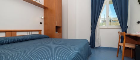 A bedroom with a bed, a wooden wardrobe, a desk with a chair, and a window with curtains.