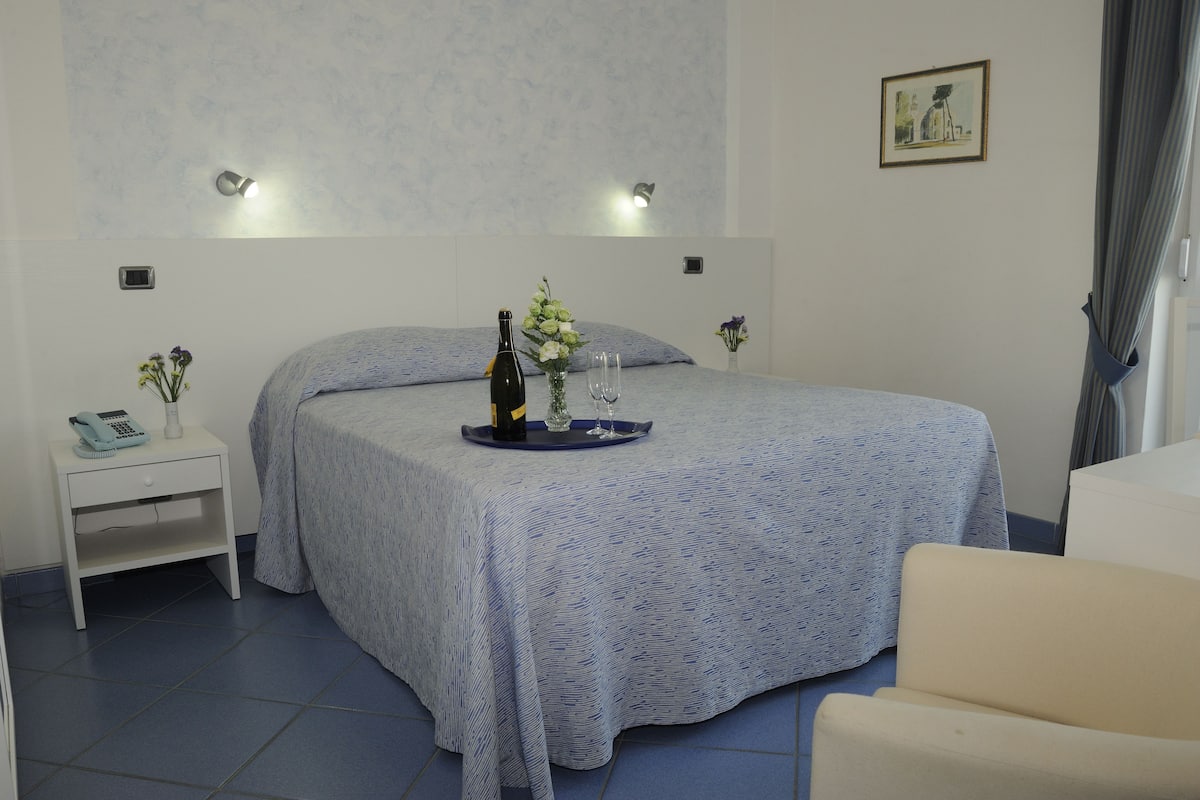 A neatly arranged hotel room with a bed, bedside tables, a chair, and a small table with a bottle and glasses.