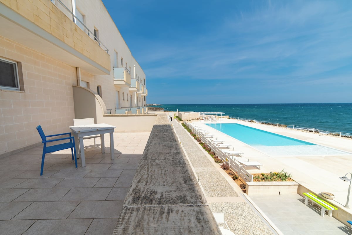 A poolside area with a view of the sea, a table, and a chair.