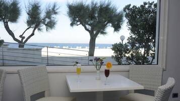 A balcony with wicker chairs and a table, overlooking a beach with a railing and trees.