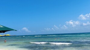 Een strand met helderblauw water, een groene parasol en enkele mensen die zwemmen.