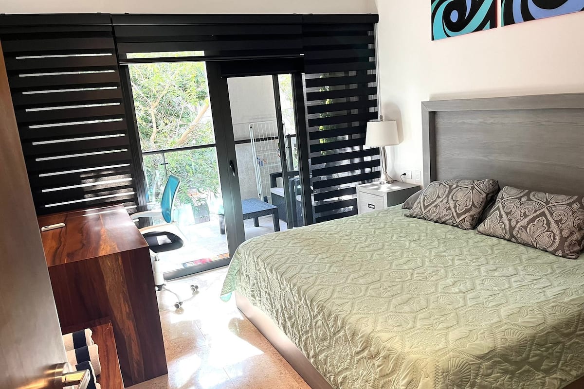 Un dormitorio con cama, escritorio, silla y balcón con vistas al verdor.