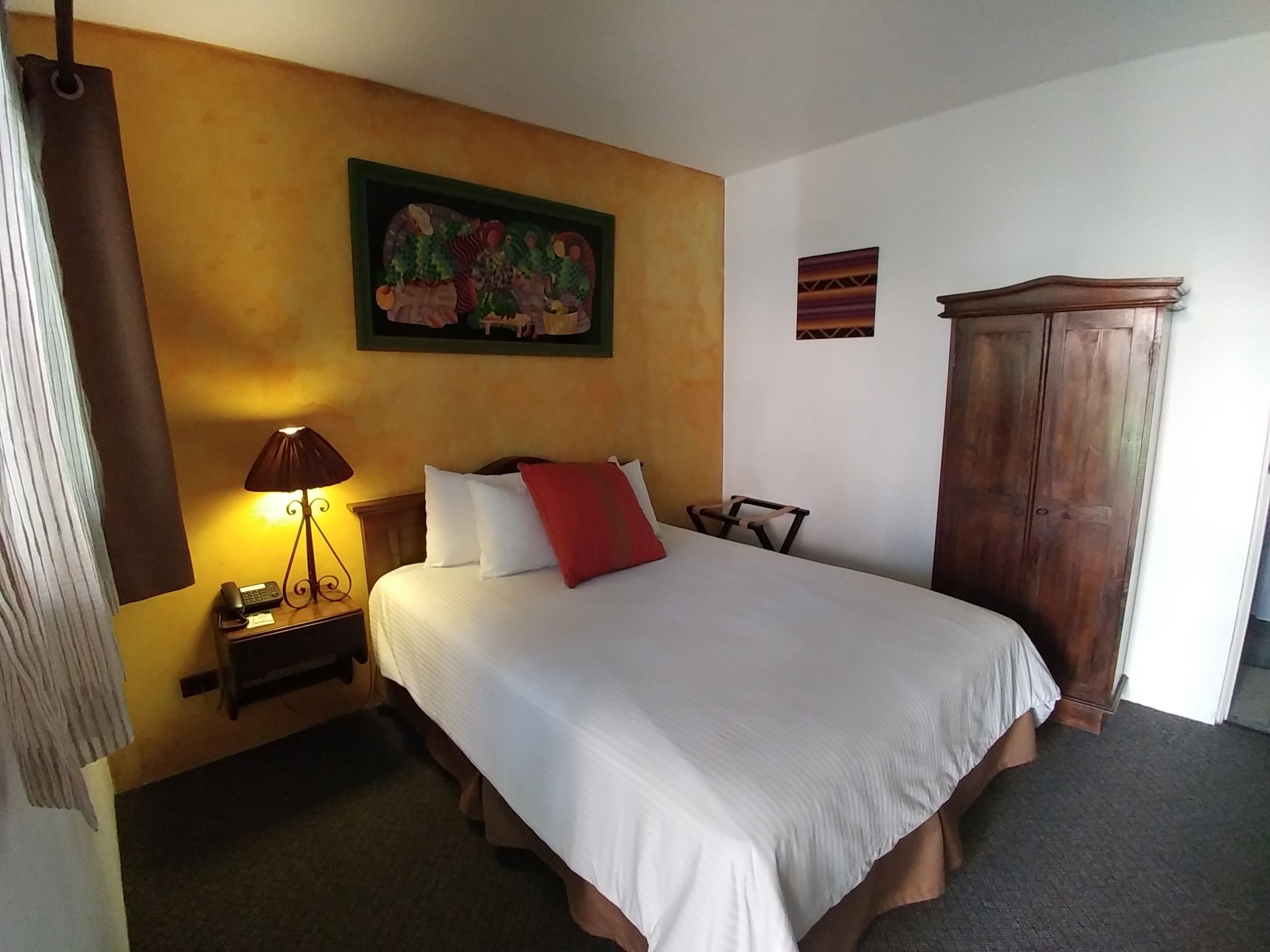 Quarto de hotel com uma cama, mesa de cabeceira com abajur, guarda-roupa de madeira e um quadro na parede.