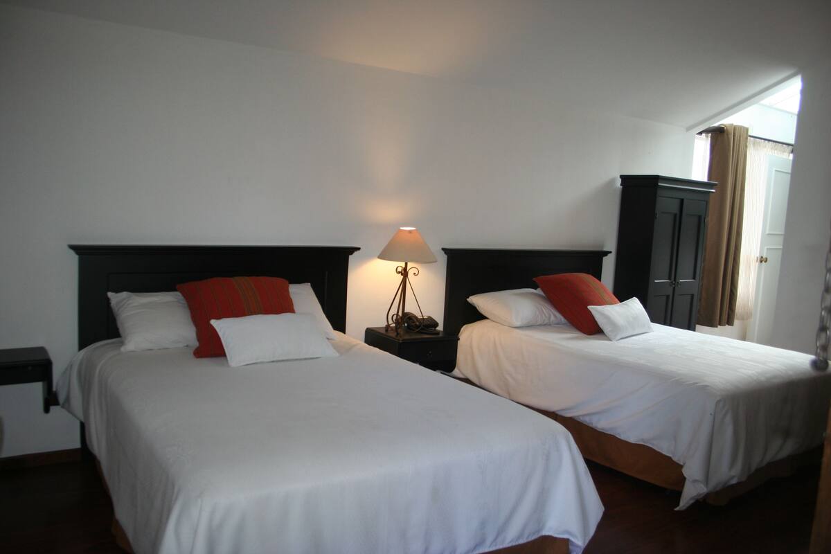 Twee enkele bedden met wit beddengoed en rode kussens, een nachtkastje met een lamp, en een donker houten garderobe.