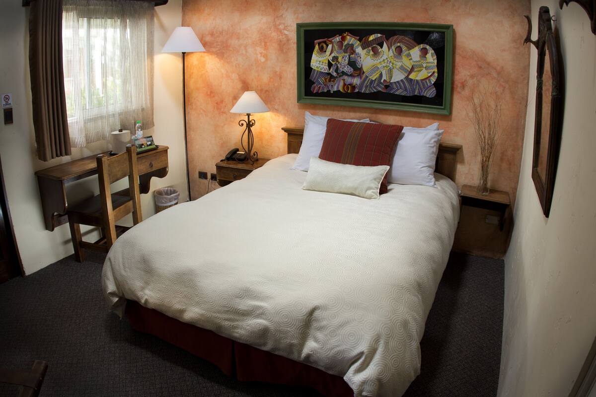 Een netjes opgemaakt bed met een bedspreide met patroon, een nachtkastje met een lamp, een stoel en een bureau met een telefoon.