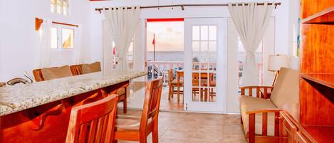 Una habitación amplia con zona de comedor, muebles de madera, ventilador de techo y vista al mar a través de una puerta.