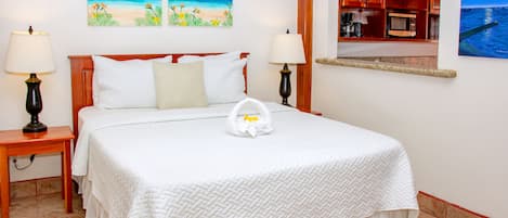 Una cama bien hecha con colcha blanca, dos lámparas de noche y una toalla con un estampado de flor amarilla. En la pared, dos cuadros que representan escenas tropicales.