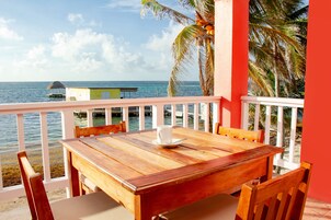Een houten tafel met stoelen op een balkon met uitzicht op de zee, en een klein gebouw en palmbomen in het zicht.