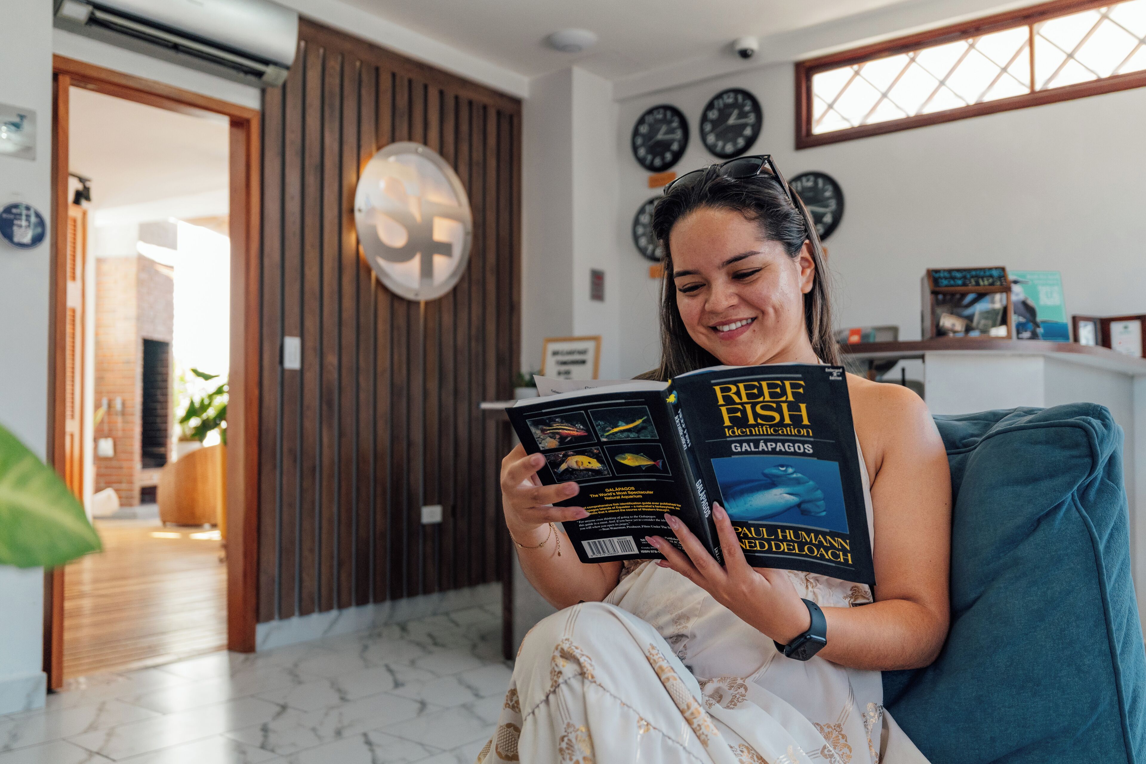 Une personne est assise sur un canapé de couleur turquoise et lit un livre intitulé « Guide d’identification des poissons de récif ».