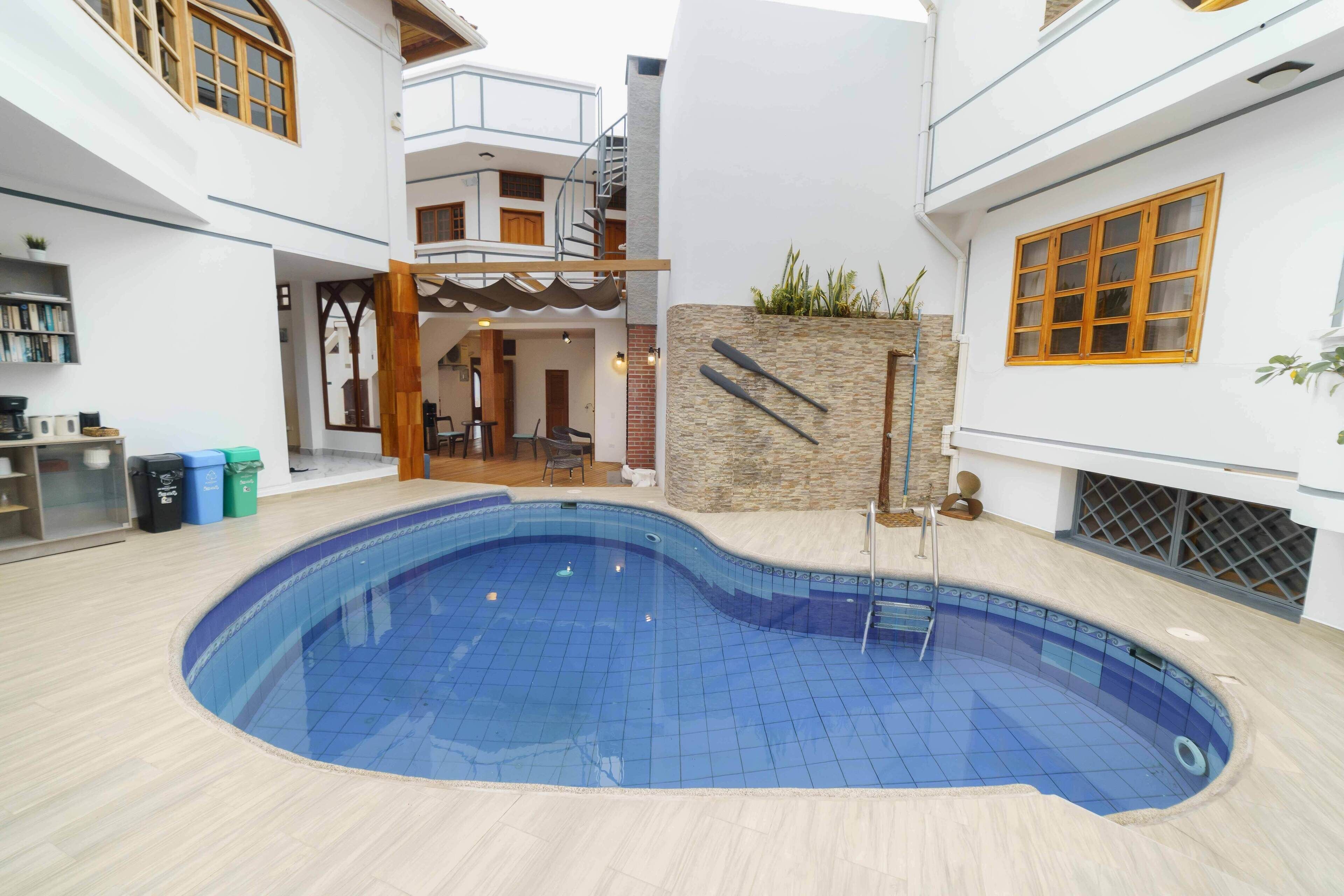 Une piscine intérieure avec un sol carrelé, entourée d’un bâtiment blanc doté d’un escalier et d’un petit jardin.