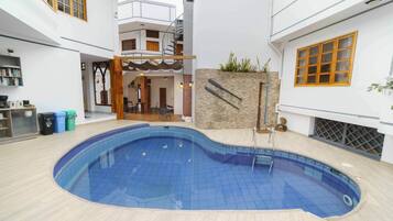 Une piscine intérieure avec un sol carrelé, entourée d’un bâtiment blanc doté d’un escalier et d’un petit jardin.
