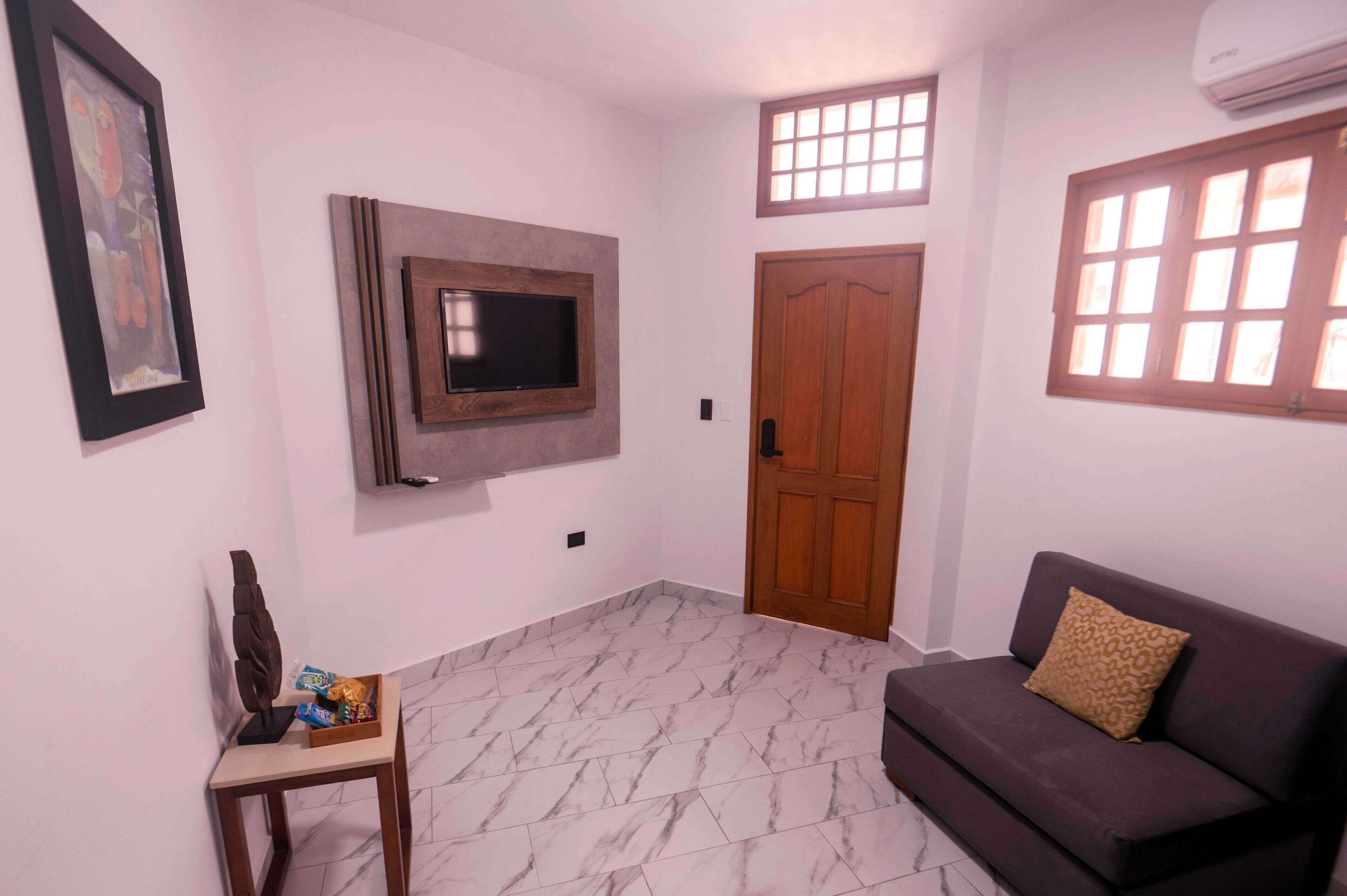 A living room with a flat-screen TV mounted on the wall, a wooden door, a window with bars, a sofa with a patterned cushion, and a small table with a decorative item and snacks.