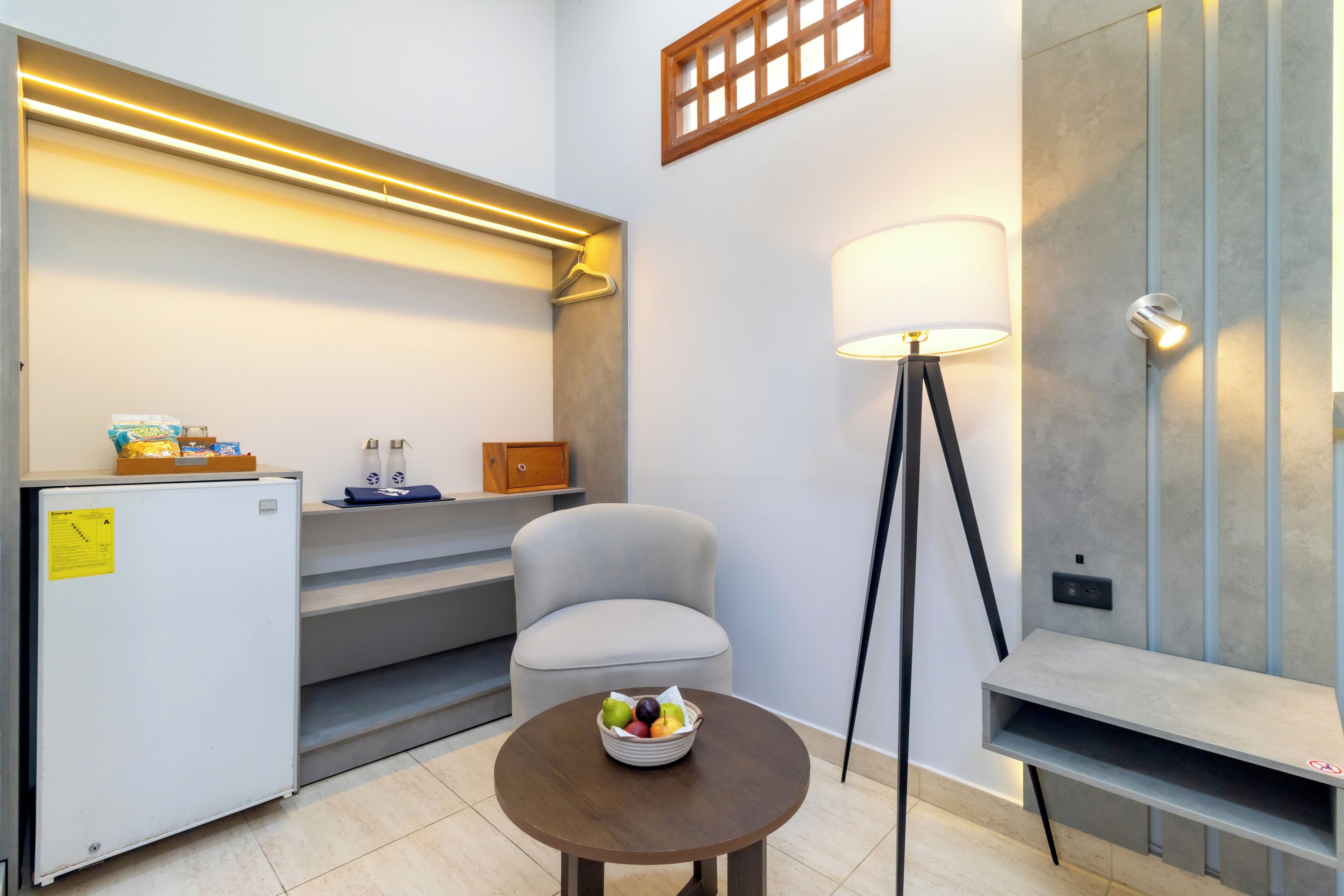 A compact hotel room with a kitchenette, a round table with fruit, a chair, a floor lamp, and a small shelf.