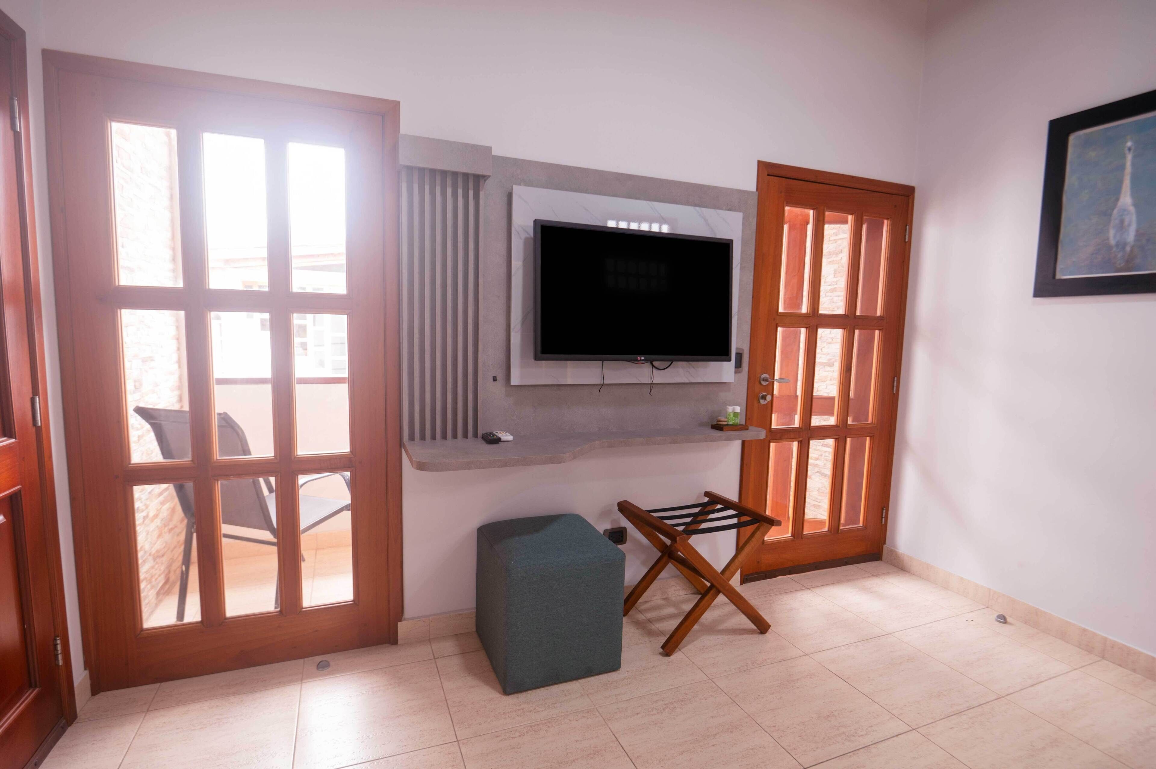 Una habitación con un ventanal grande, un televisor de pantalla plana montado en la pared, una mesita pequeña y una silla plegable.