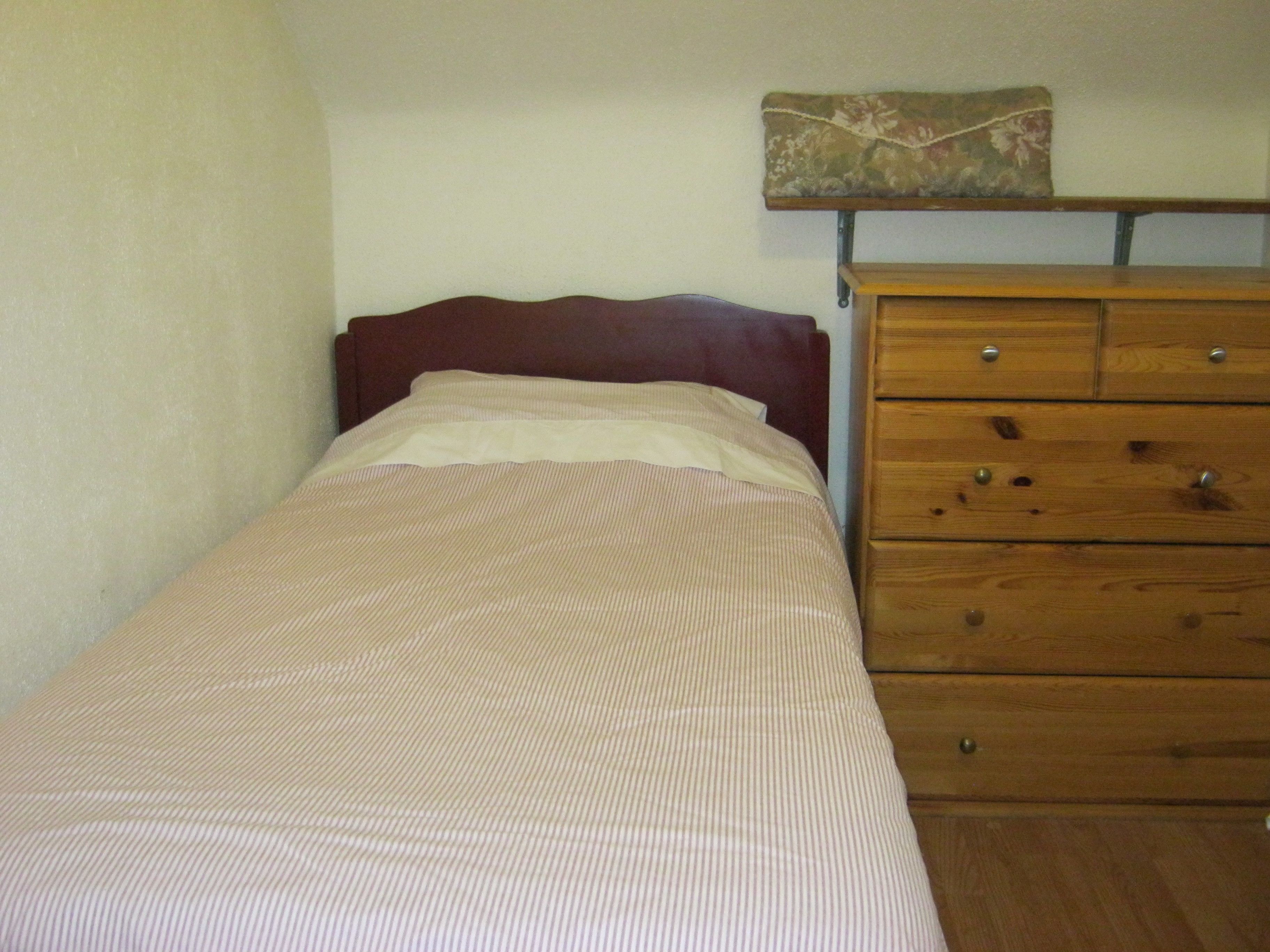 A small, single-bed room with a wooden dresser and a floral-patterned pillow on a shelf above the bed.