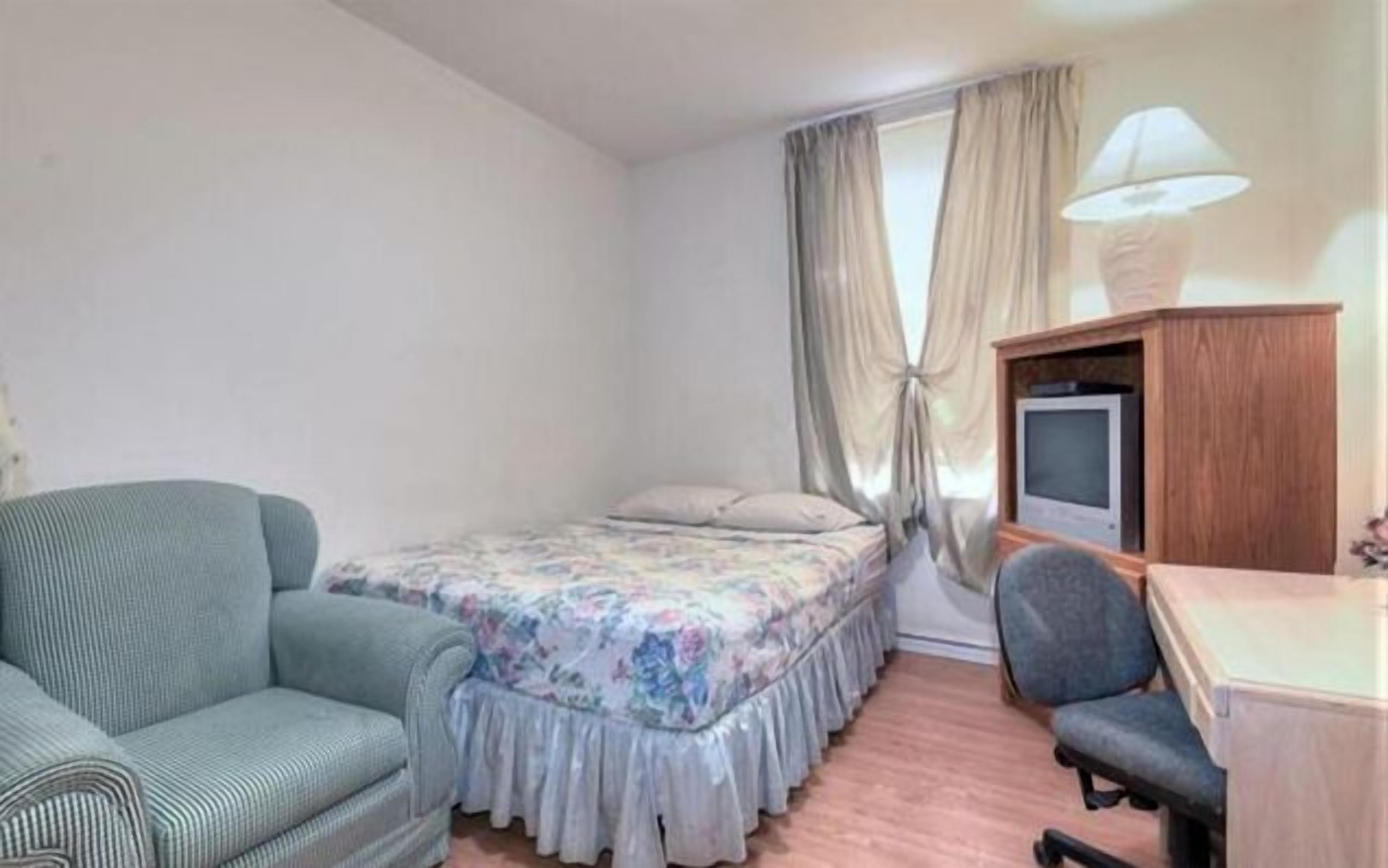 Una habitación pequeña y ordenada con cama, una silla, escritorio y televisor.