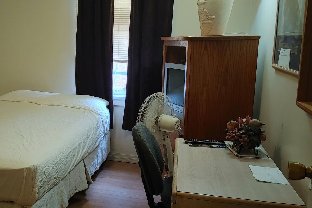 Un dormitorio con una cama individual, una silla, un escritorio con un ventilador y un armario.