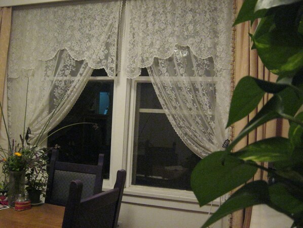 Una habitación con una ventana cubierta por cortinas de encaje blanco, una mesa de madera con un jarrón de flores y una silla.