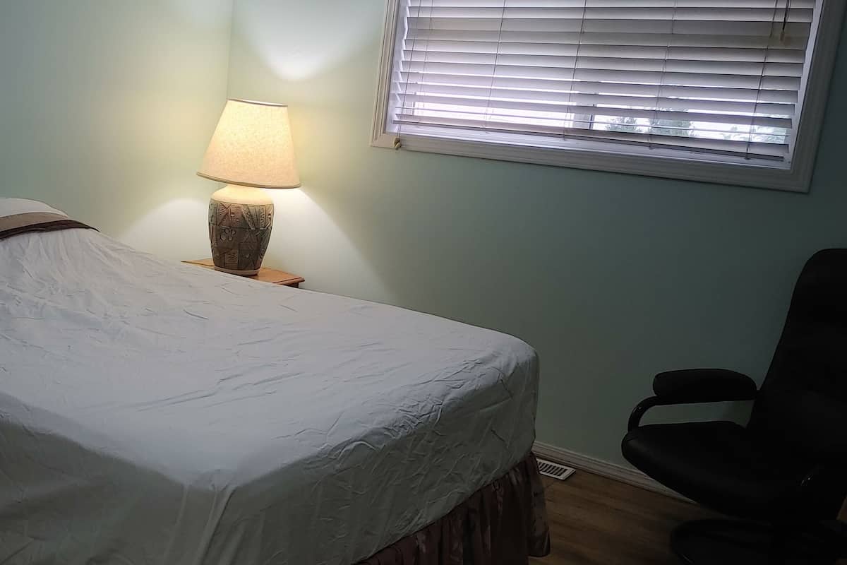 Una cama bien hecha con una colcha blanca y una manta marrón, una lámpara de noche sobre una mesita de madera, una ventana con persianas y una silla negra a la derecha.