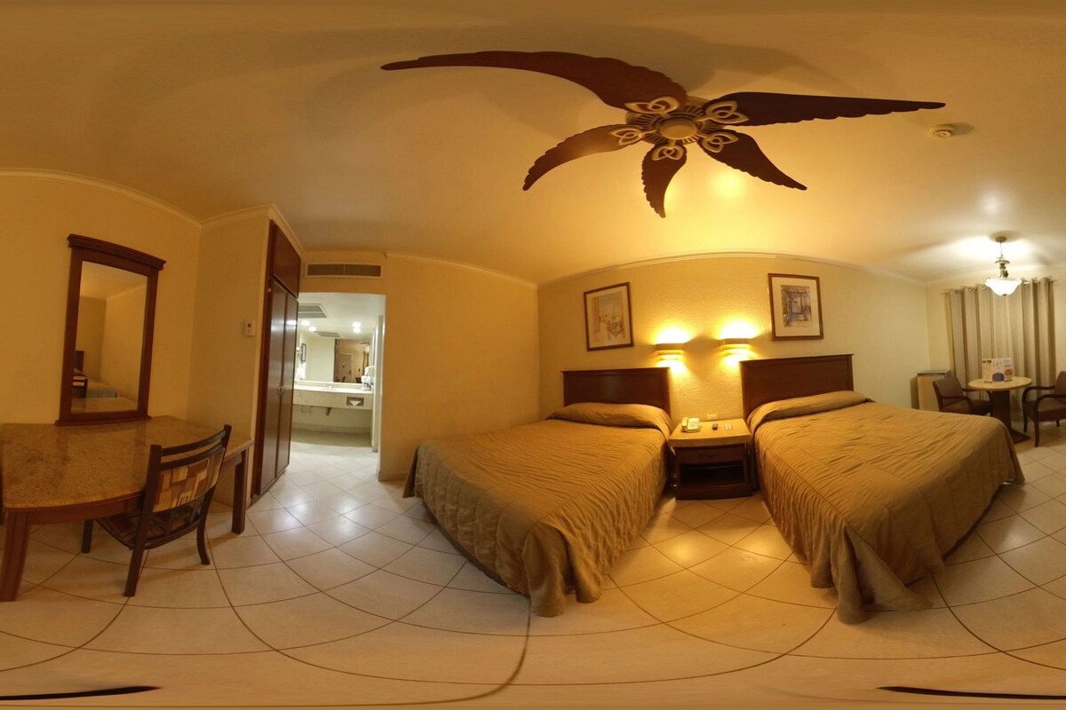Una habitación de hotel con cama, escritorio, silla, televisión y ventilador de techo.