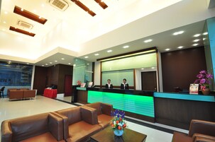 Zona de recepción de hotel con diseño moderno, que incluye un mostrador, dos recepcionistas, un área de descanso con sofás de cuero y un jarrón de flores sobre una mesa.