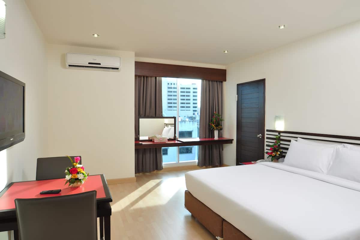 A hotel room with a bed, a desk with a chair, a flat-screen TV, an air conditioning unit, a window with curtains, and a view of buildings outside.