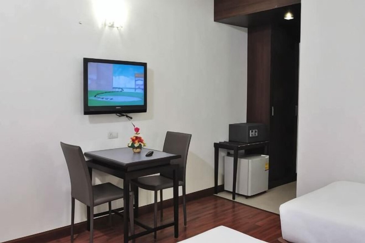 A small room with a TV mounted on the wall, a table with chairs, a white sofa, and a small refrigerator.
