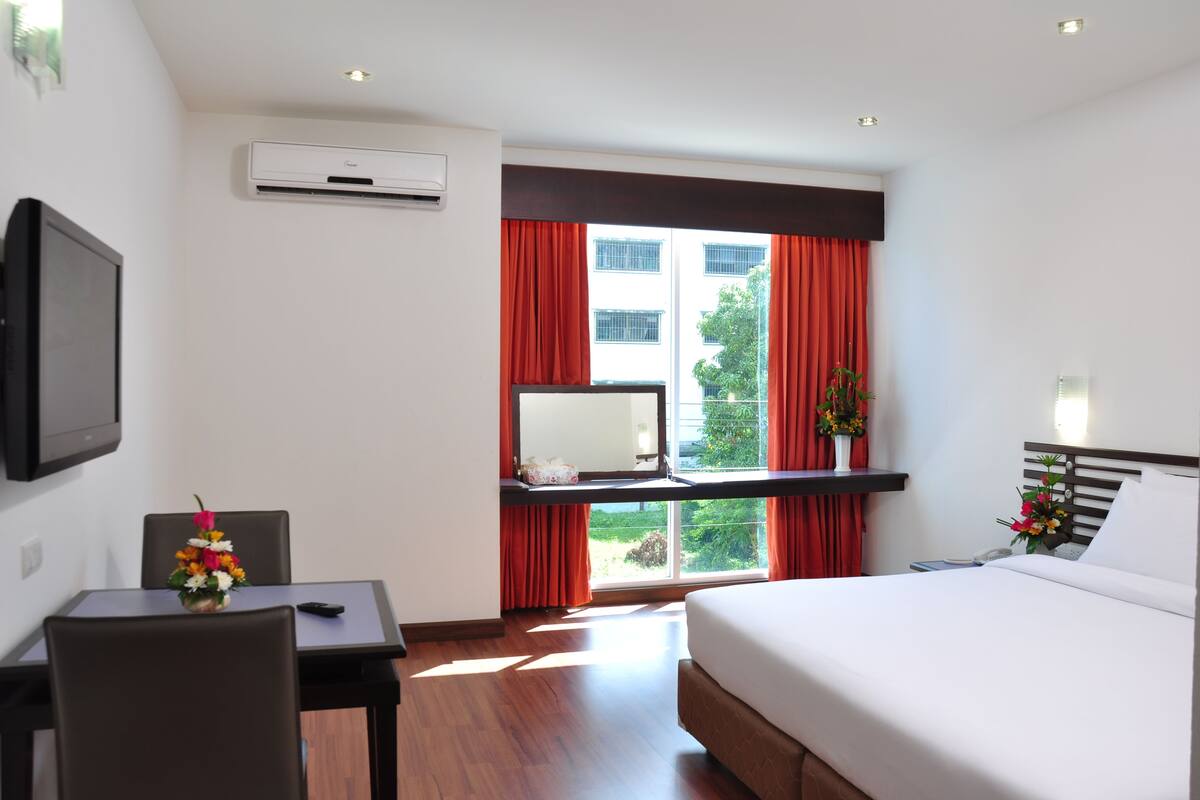 A hotel room with a bed, a desk with a chair, a TV mounted on the wall, and a window with red curtains.