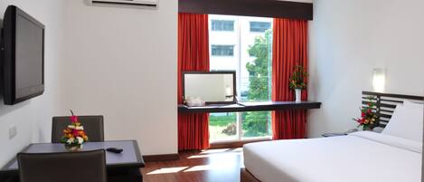 A hotel room with a bed, a desk with a chair, a TV mounted on the wall, and a window with red curtains.
