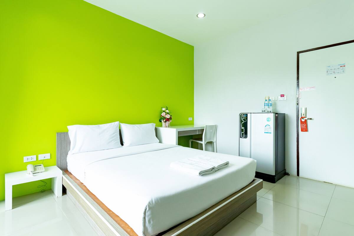 A modern hotel room with a bright green accent wall, a neatly made bed, a small desk with a chair, a refrigerator, and a white bedside table with a phone.