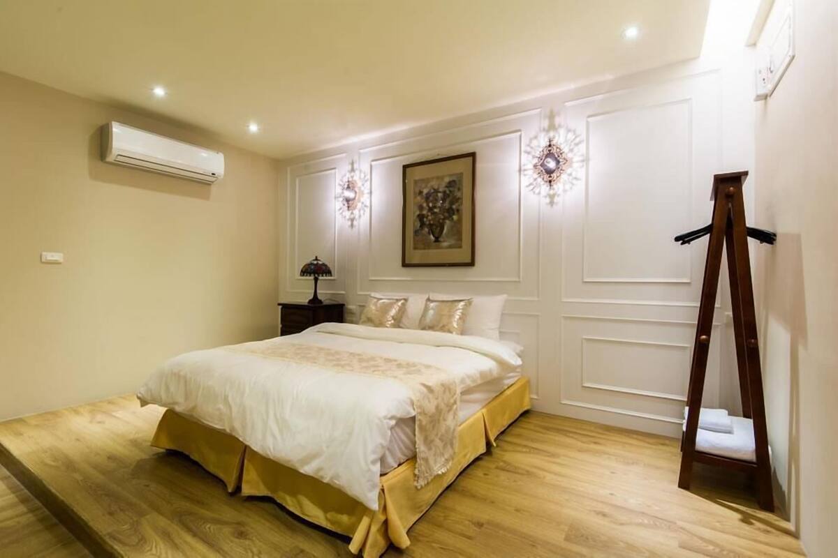 A bedroom with a large bed, an air conditioner, a nightstand, a lamp, a painting, and a wooden ladder.