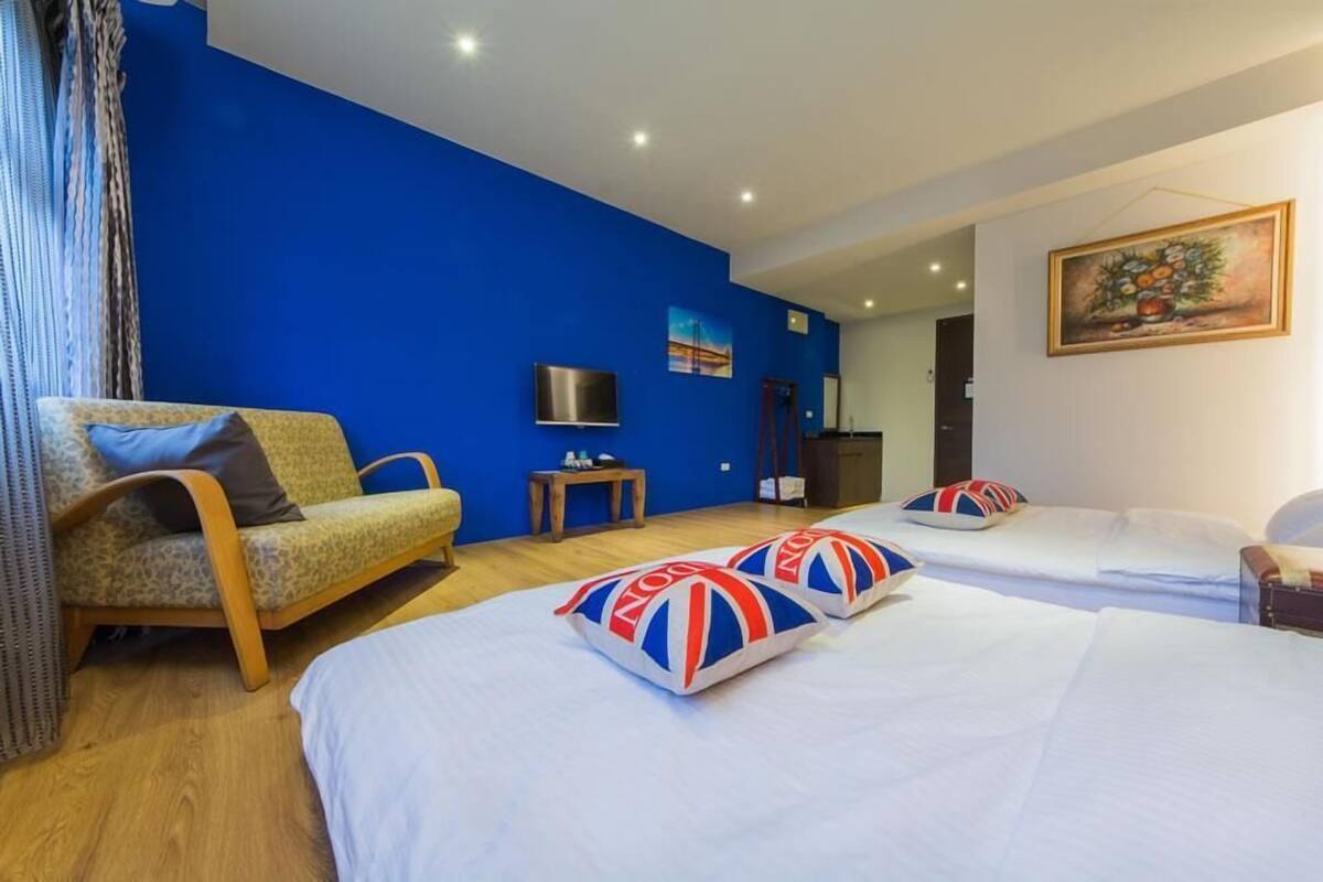 A hotel room with a blue accent wall, a sofa, two beds with matching pillows, a TV, and a painting.